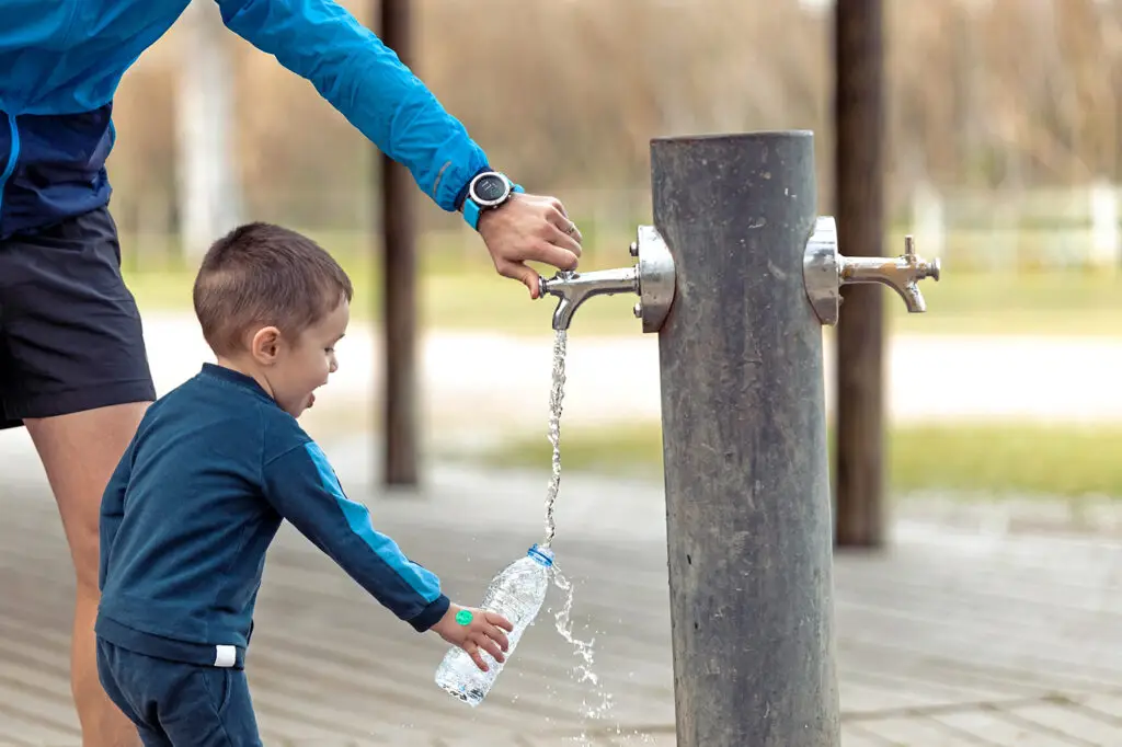 Funny little boy filling his water bottle in public fountain whi